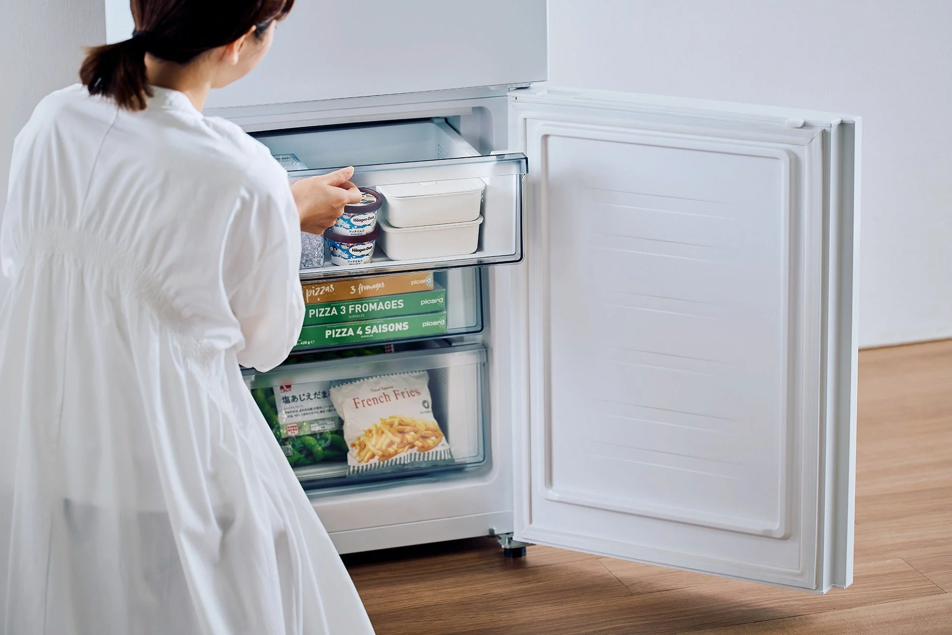 白い冷蔵庫の冷凍室を開け、中に保存されている冷凍食品を取り出している女性の様子。ハーゲンダッツのアイスクリームやピザ、フライドポテトなどがきれいに収納されている。