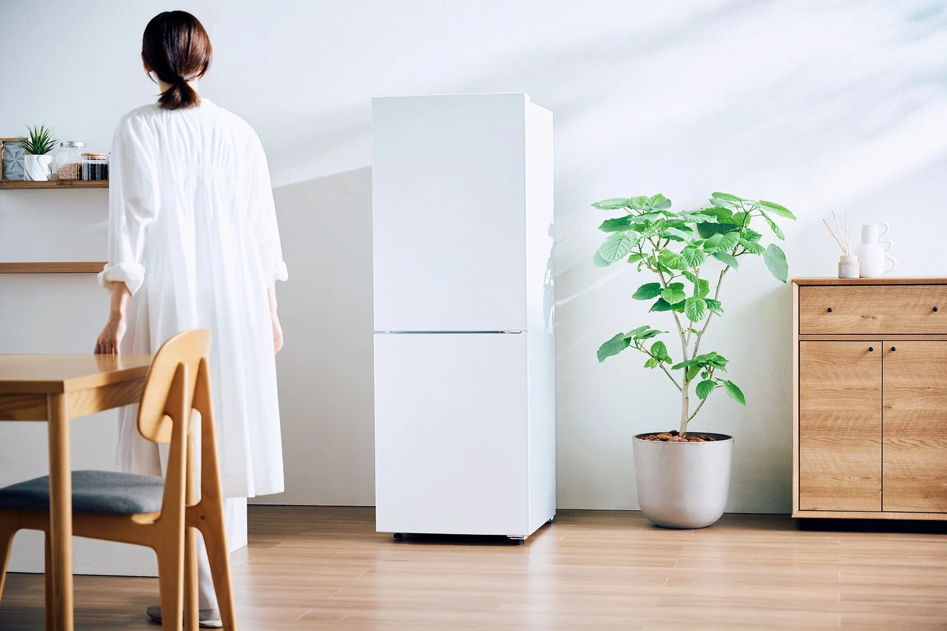白い壁と木目調の家具で統一された清潔感のある部屋で、女性が冷蔵庫のそばに立っている後ろ姿を捉えた一枚。観葉植物が配置され、明るく落ち着いた生活空間が表現されています。