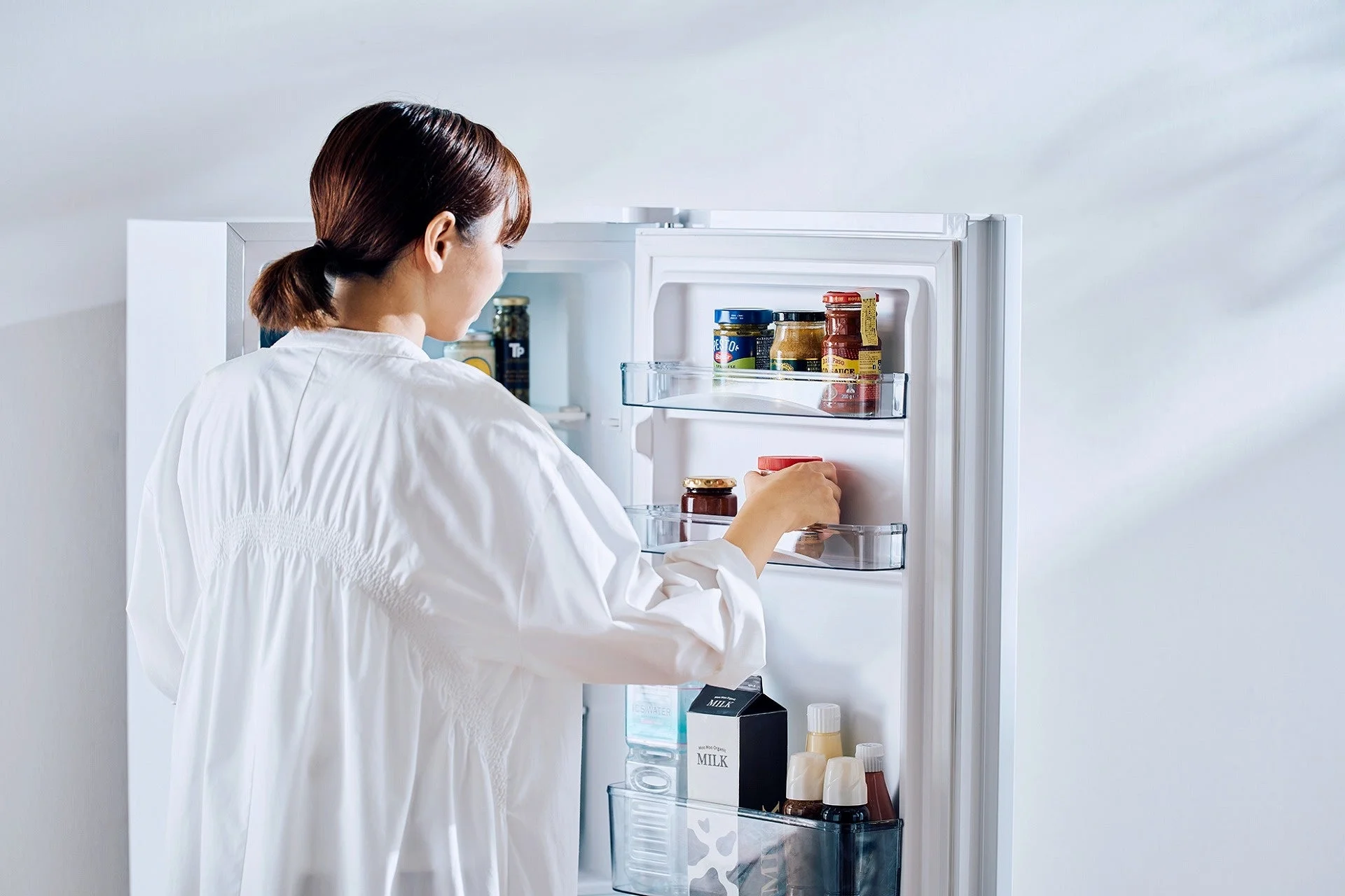 白いシャツを着た女性が、開いた冷蔵庫の中の食品を整理している様子です。冷蔵庫には牛乳パックや様々な瓶詰めの食品が整然と収納されています。