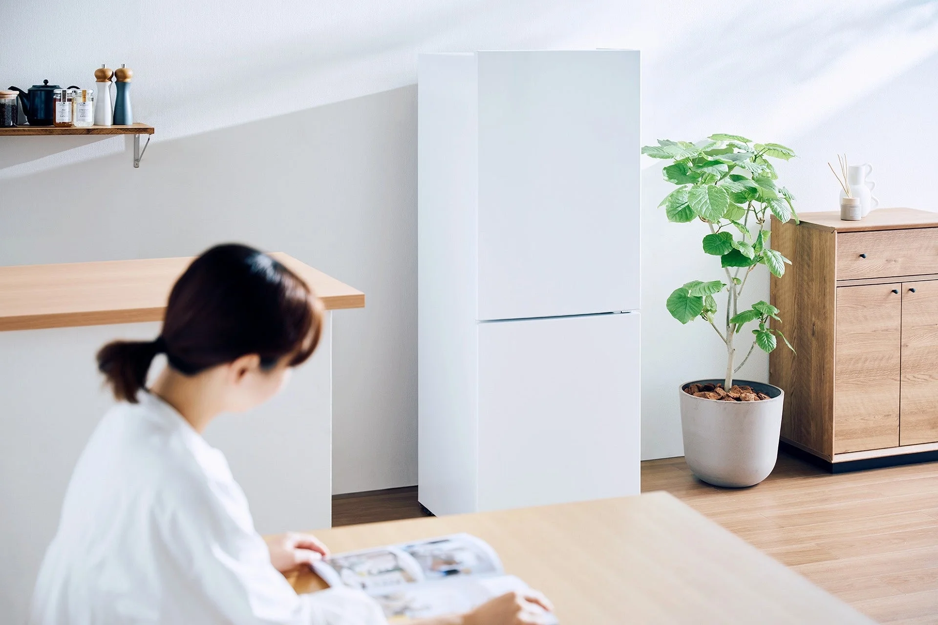 明るいリビングまたはダイニングルームで、一人の女性が木製のテーブルで雑誌を読んでいます。背景には白い冷蔵庫、大きな観葉植物、木目調の収納棚があり、シンプルで落ち着いた雰囲気です。