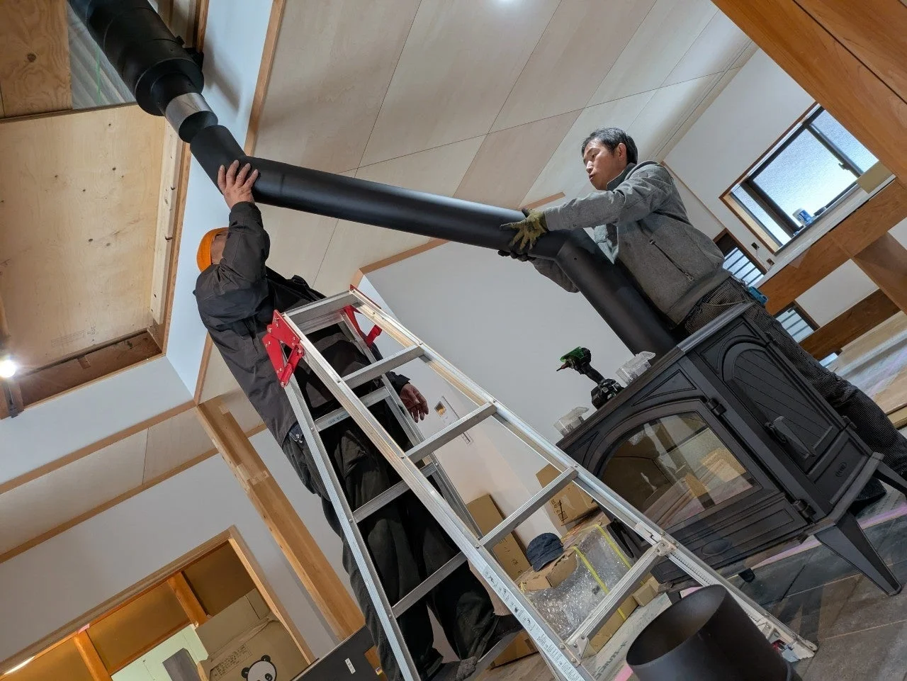 二人の作業員が室内で薪ストーブに黒い煙突を設置している様子