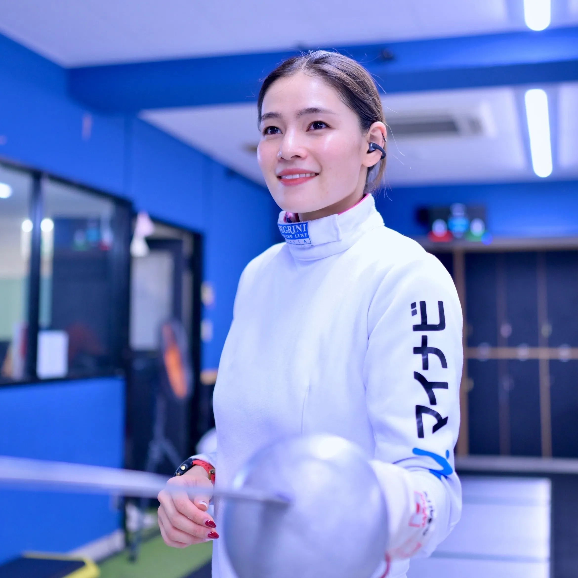 フェンシングの防具を身につけた女性が、フェンシングの剣を持ち、笑顔でこちらを見ている様子