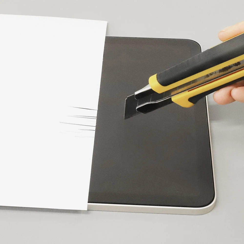 カッターナイフの刃で紙を切りながら、その下のタブレットの画面に傷をつけようとしているように見える