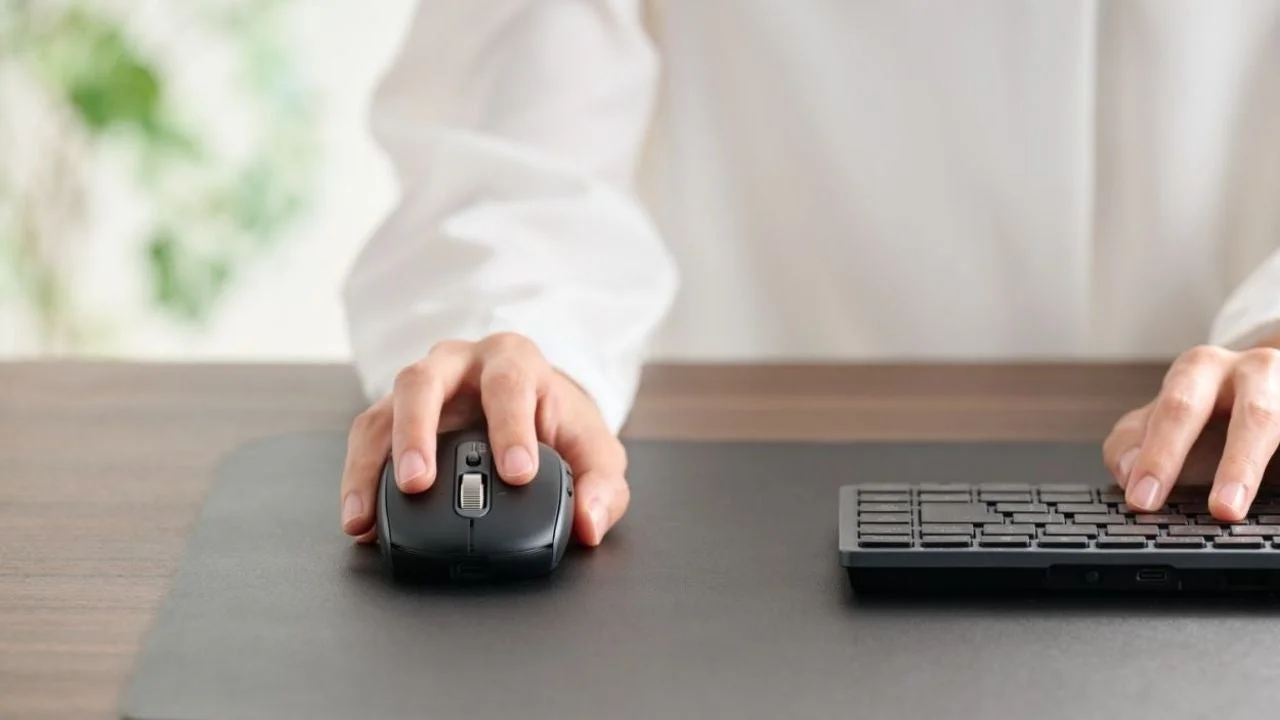 デスクでパソコンのマウスとキーボードを操作している手元のクローズアップ画像です。白いシャツを着た人物が、黒いマウスパッドの上で作業しています。