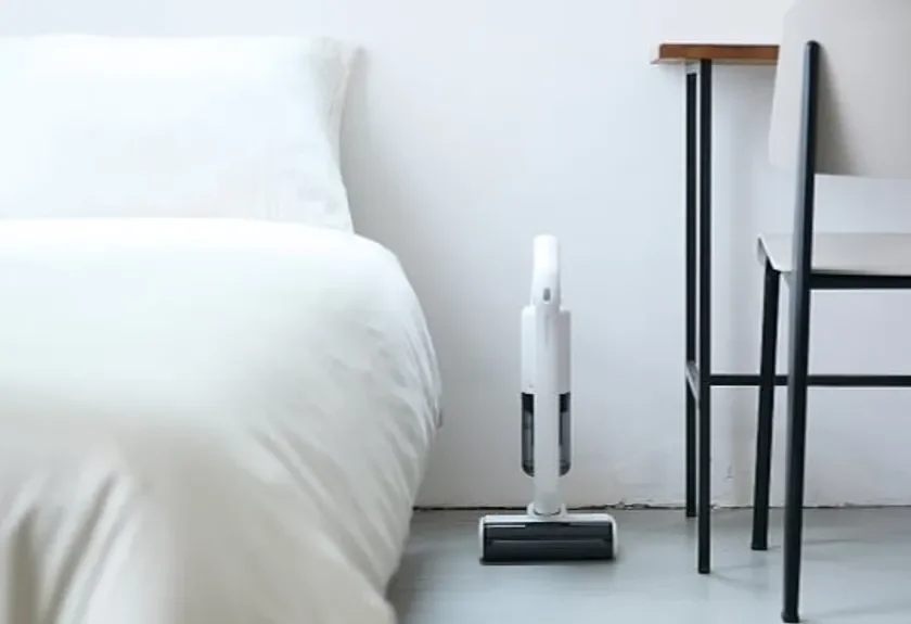 白い布団が敷かれたベッドの横に、白いコードレス掃除機が立てて置かれています。