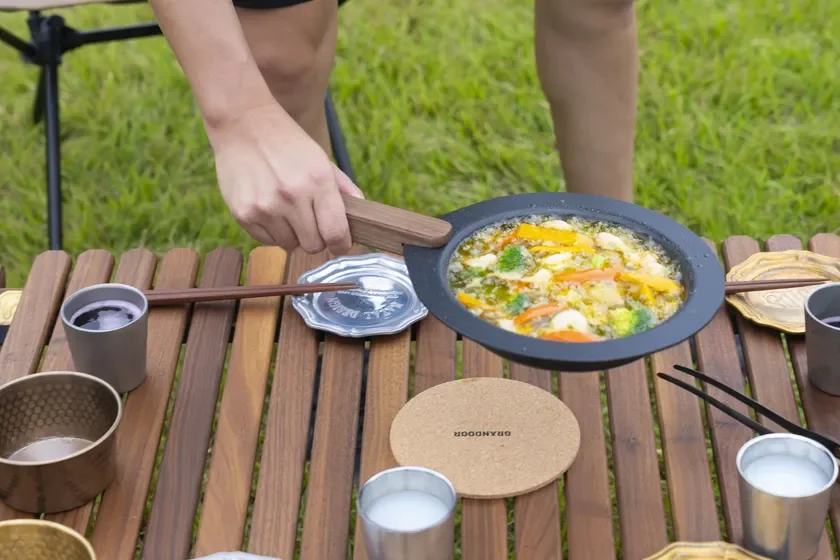 屋外の木製テーブルで、野菜がたっぷり入った鍋料理を準備している様子