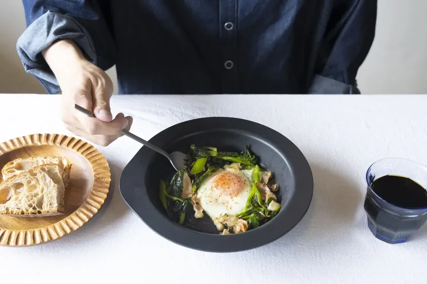 白いテーブルクロスの上で、デニムシャツを着た人が目玉焼きと緑の野菜が入った黒いボウル、パン、コーヒーの朝食を食べています