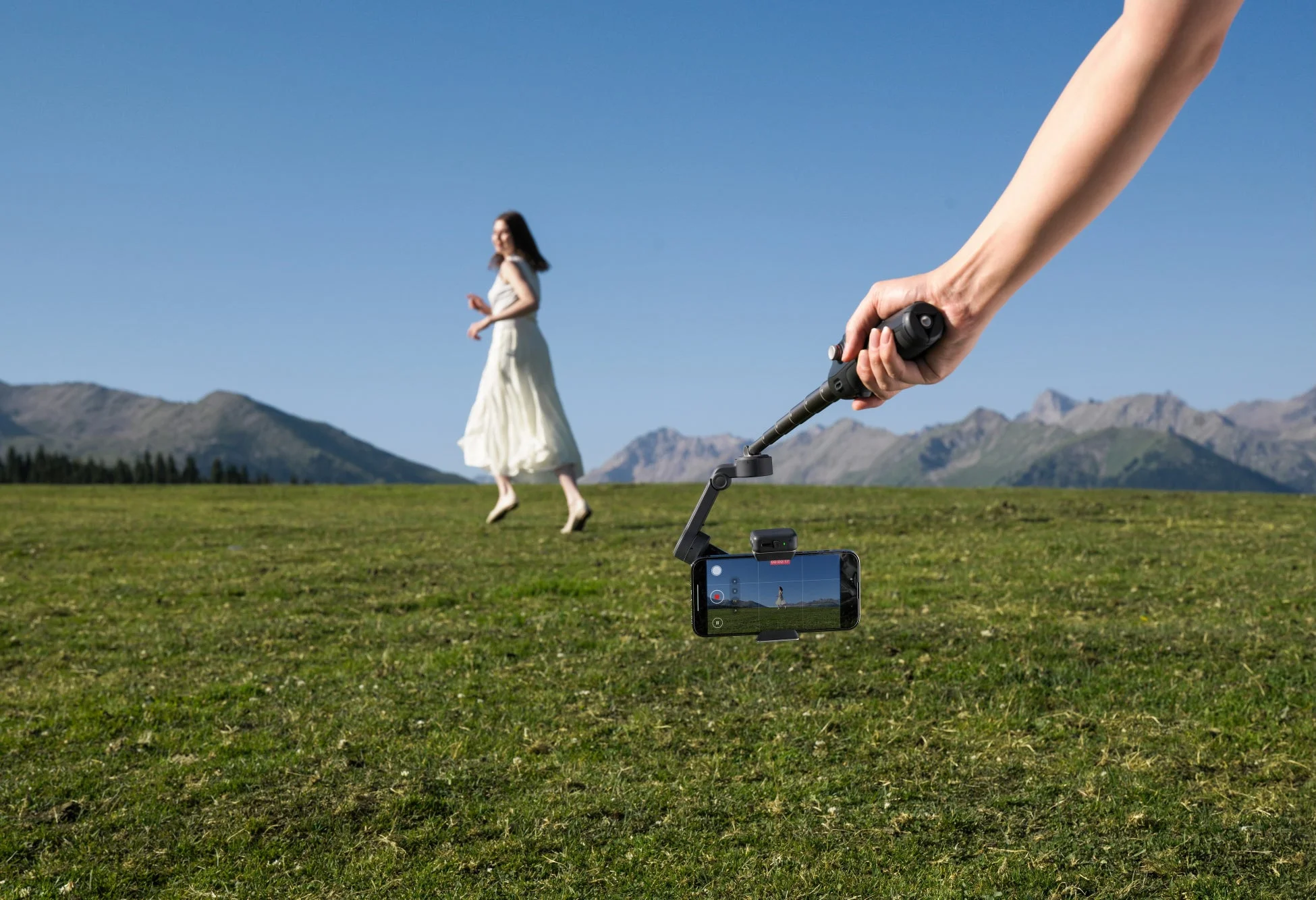 広大な草原でスマートフォンとジンバルを使い、白いドレスの女性が歩く姿を撮影しているシーン