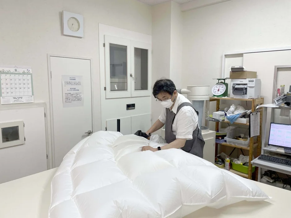 白い作業着を着た人が、清潔な作業台の上で大きな羽毛布団を丁寧に確認している