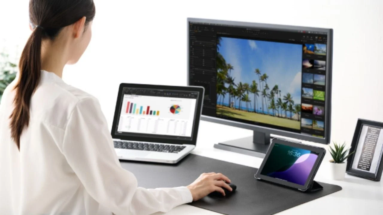 白いシャツを着た女性が、ノートパソコンと大型モニター、タブレットが並ぶデスクで作業しています。現代的なオフィスやリモートワークの風景を表しています。