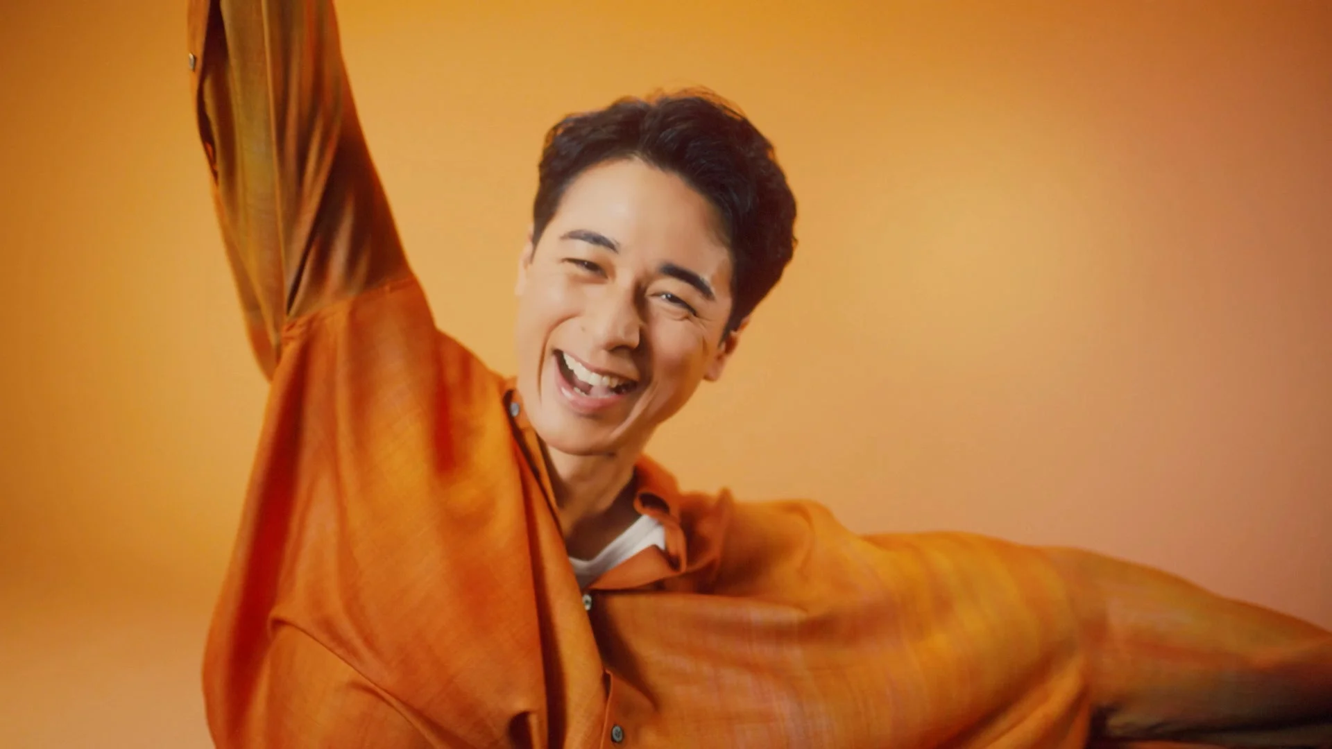 オレンジ色のシャツを着た男性が、明るい背景の前で満面の笑み。