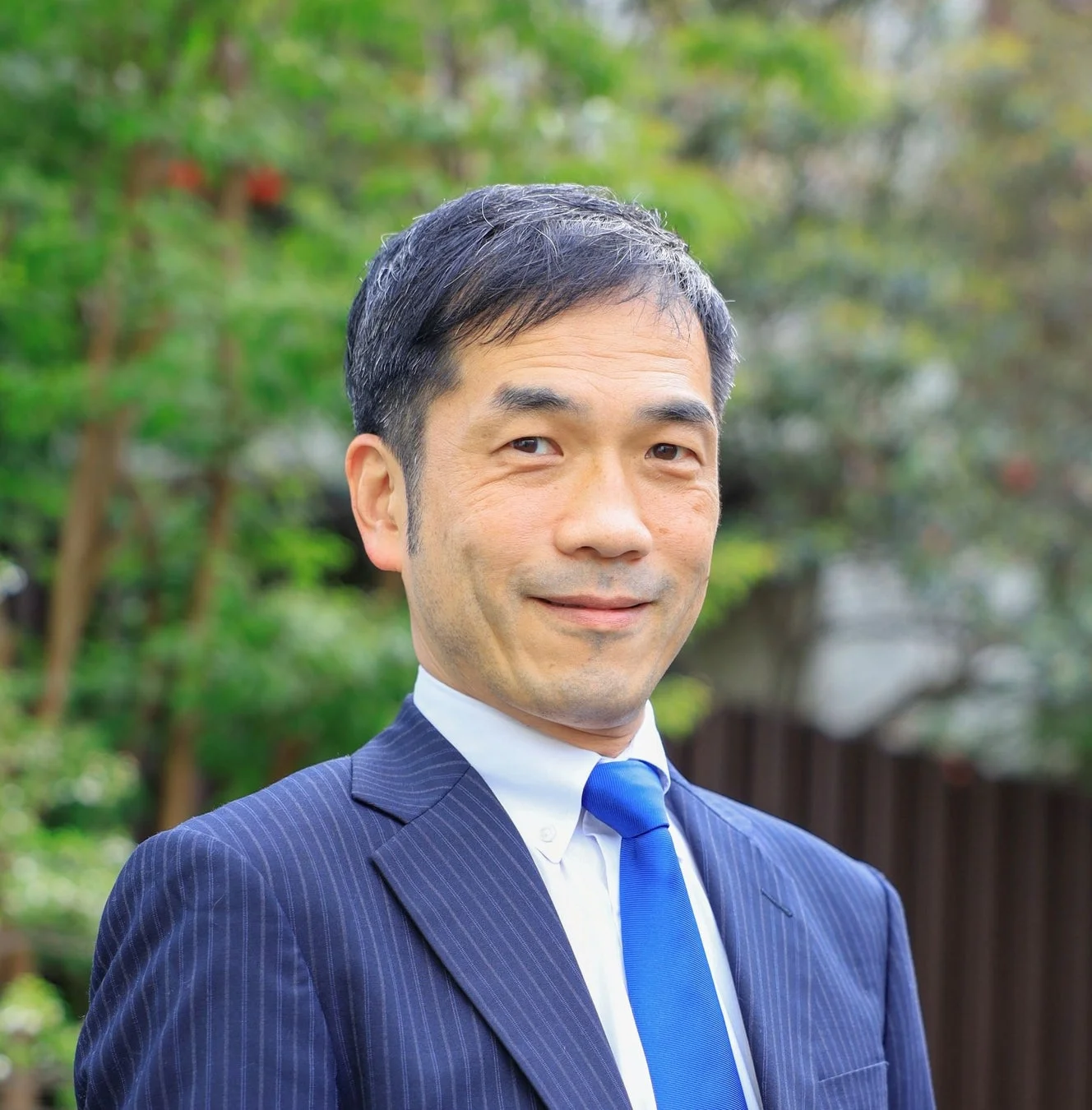 青いネクタイとストライプのスーツを着用したアジア系男性が、屋外の緑を背景に穏やかに微笑むポートレートです。