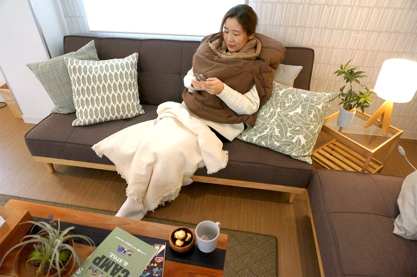 リビングのソファで女性がくつろいでいます。茶色の着る毛布に包まれ、スマートフォンを操作しています。クッションや観葉植物があり、暖かく居心地の良い空間です。