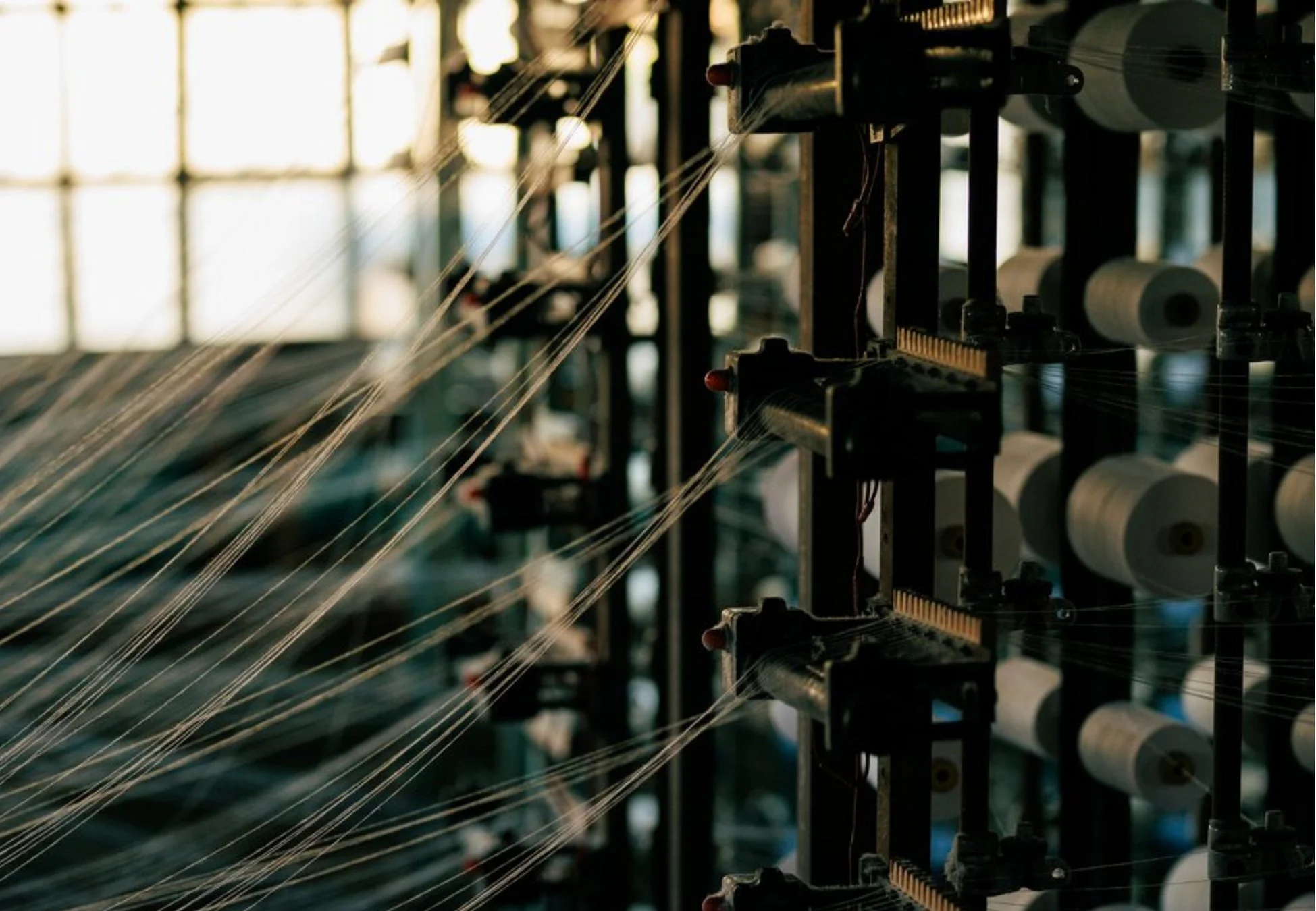 織物工場の機械で多数の白い糸が張られ、糸巻きが並ぶ様子