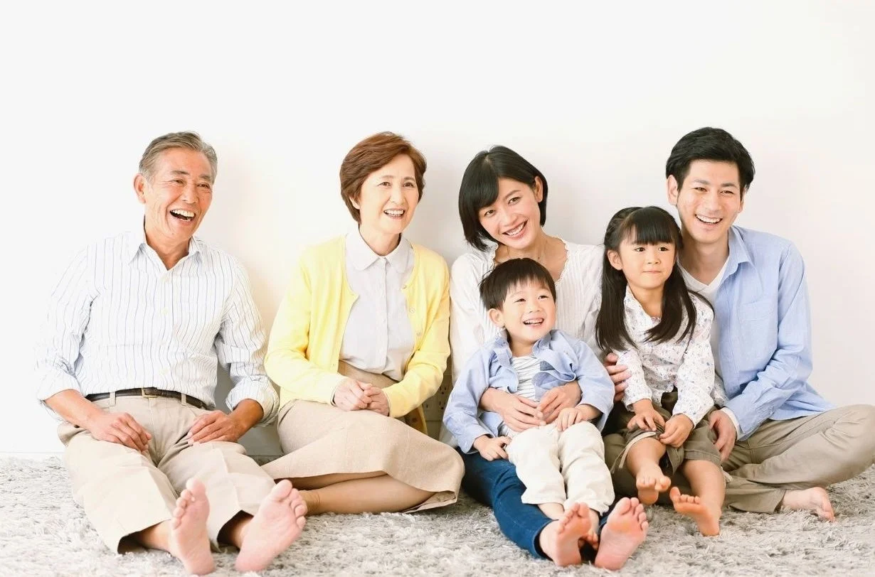 白い壁を背景に、三世代の家族6人が笑顔でカーペットに座っている様子。祖父母、両親、2人の子供たちが幸せそうな表情を浮かべている。