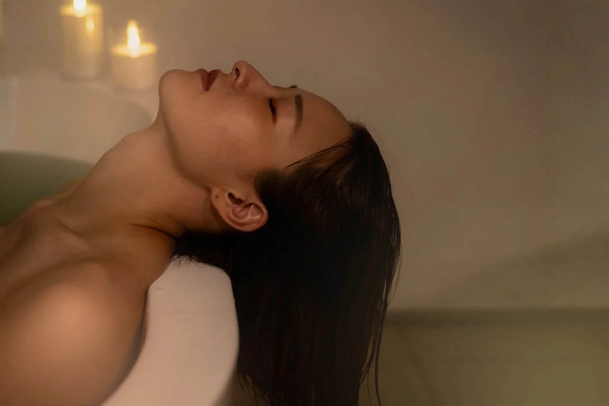 キャンドルの光が灯るバスルームで、女性が湯船に浸かりリラックスしている様子。