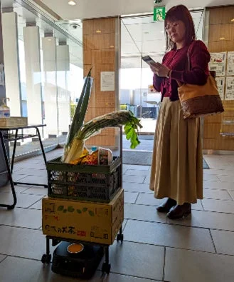女性がスマートフォンを操作し、食料品を運ぶ自律型配送ロボットから荷物を受け取ろうとしている様子
