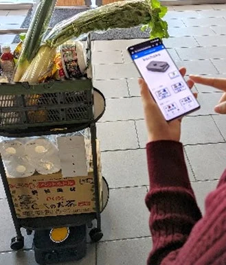スマートフォンで配送アプリを操作している人物と、野菜や日用品が積まれたデリバリーカートの様子