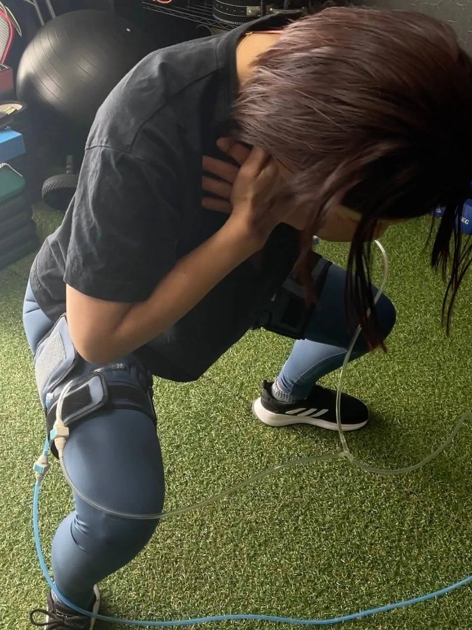 人工芝の上で、特殊な運動器具を装着した女性がスクワットのような体勢でトレーニングをしています。体勢から運動の負荷や集中がうかがえ、ジムのような環境で健康維持や体力向上に取り組んでいる様子です。