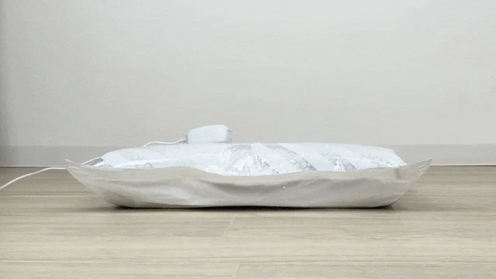 白い壁と木目調の床を背景に、真空圧縮袋が置かれています。袋の中の衣類や布団が、白い電動ポンプによって効率的に圧縮されている様子です。収納スペースを有効活用するための実用的なアイテムの使用例を示しています。