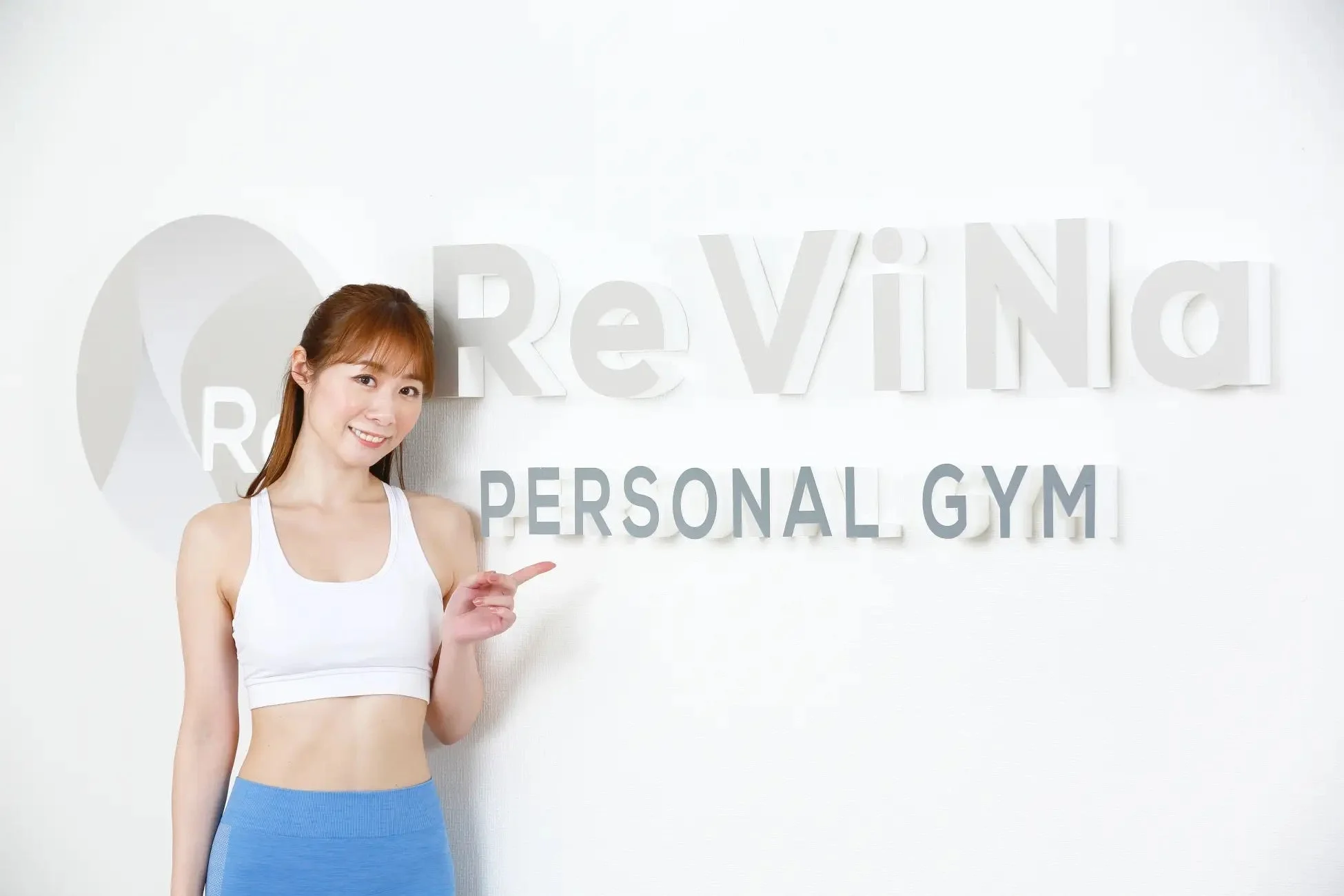 ReViNa PERSONAL GYMのロゴを背景に指を指す女性