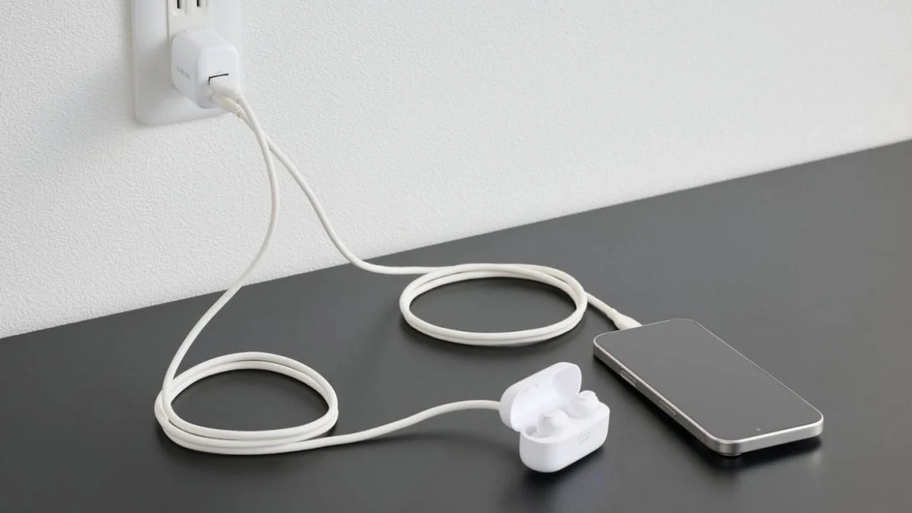 壁のコンセントに差された白いUSB充電器が、長いケーブルでスマートフォンとワイヤレスイヤホンケースに接続され、同時に充電している様子。黒いデスク上でデジタル機器が並べられている。