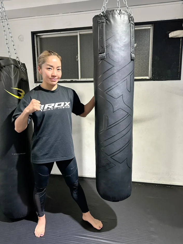 スポーツウェアを着た女性がジムでサンドバッグの横に立ってポーズをとっています。