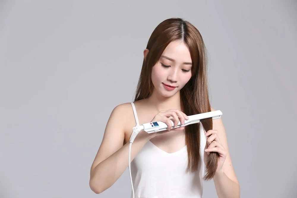 白いタンクトップを着た女性が、白いヘアアイロンを使って自身の長い髪をスタイリングしている。背景はシンプルなグレー。