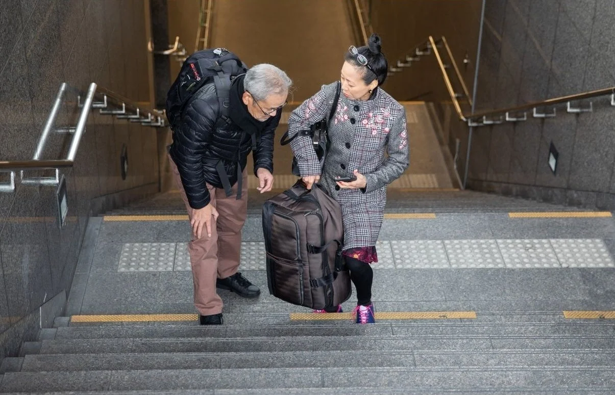 地下通路の階段で、高齢の男女が重そうな荷物を運びながら移動している様子。男性が女性の荷物を持つ手伝いをしている。