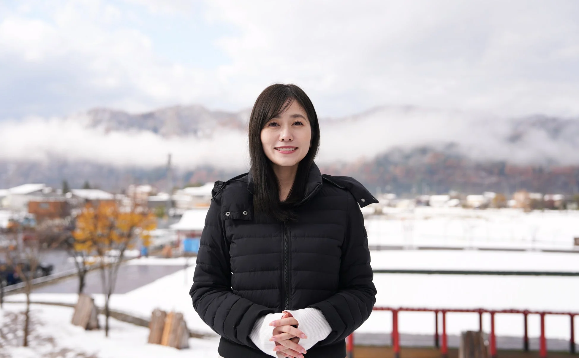 雪が積もった景色を背景に、黒いダウンジャケットを着た女性がカメラに向かって微笑んでいます。