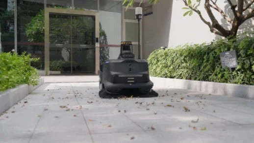 屋外の舗装された通路で、黒い自律型清掃ロボットが落ち葉を掃いています。