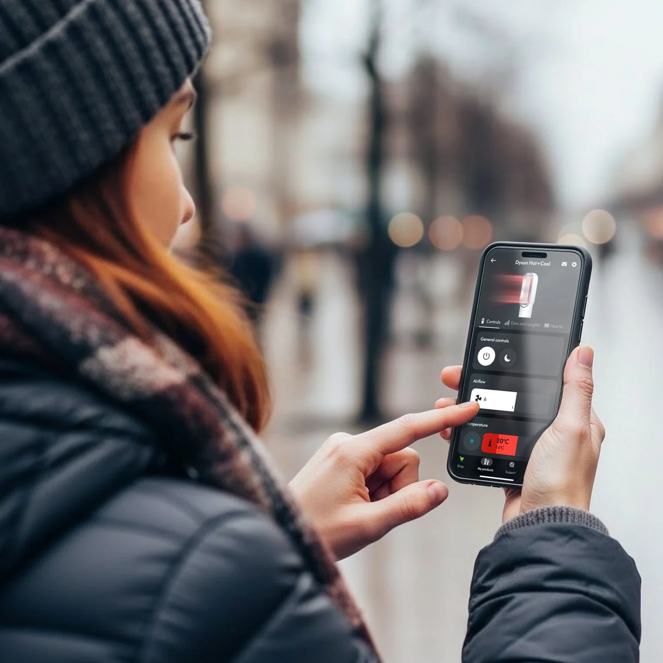 冬の屋外で、女性がスマートフォンを操作しDyson Hot+Coolのアプリ画面を見ている。気温や風量を設定しており、スマート家電を遠隔操作している場面。