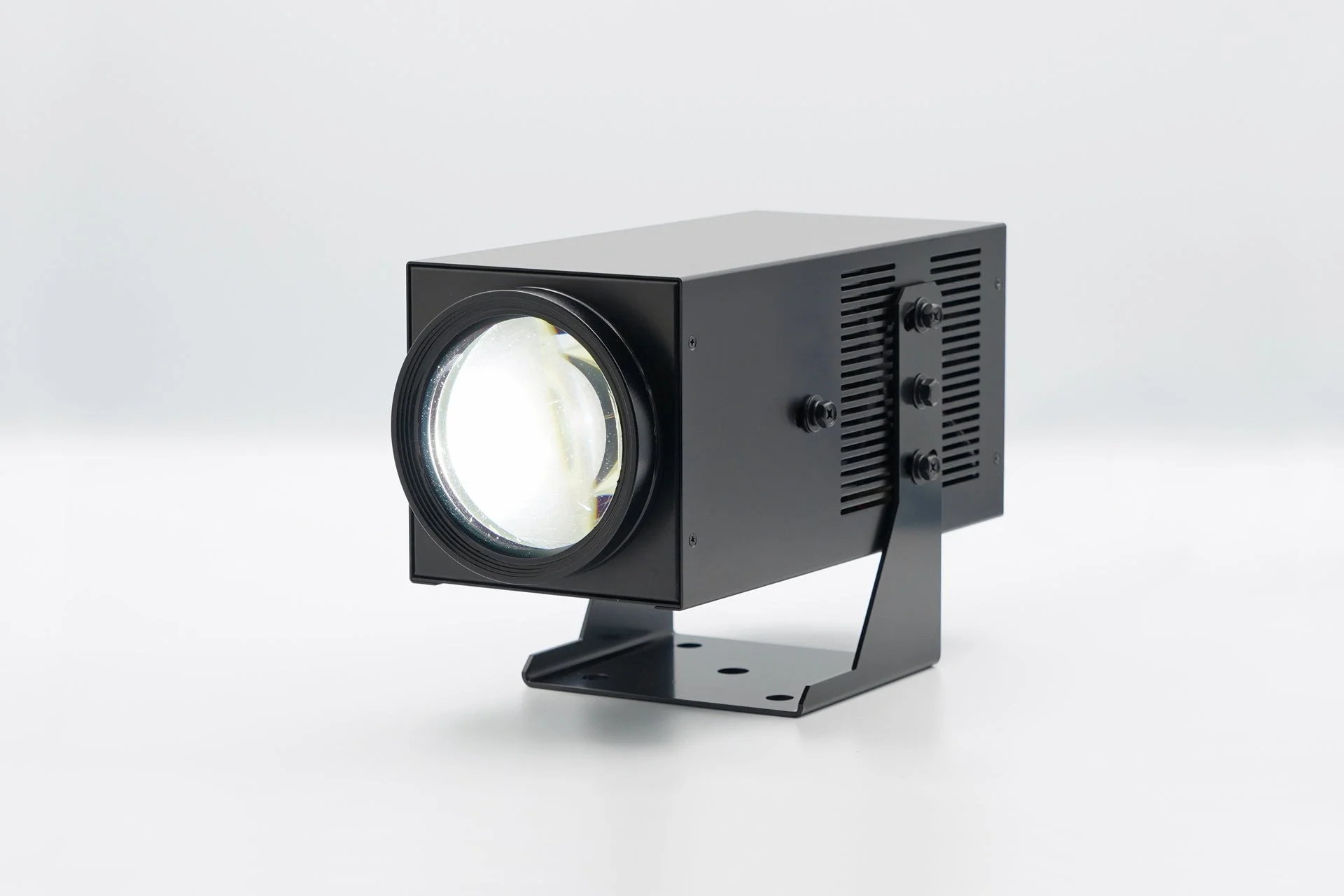 白い背景に置かれた黒い箱型プロジェクターのような照明器具のクローズアップ画像です。前面のレンズ部分から明るい光が放たれています。