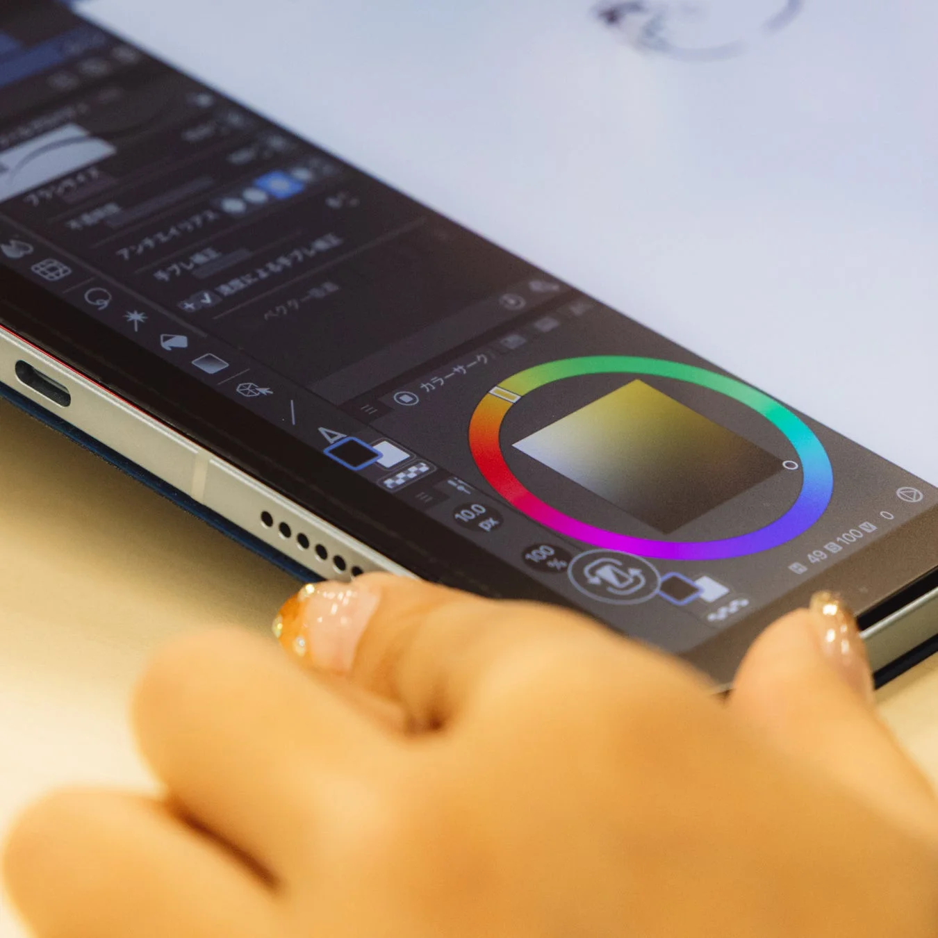 タブレットでデジタルアートを作成している様子が写っています。画面には描画アプリのインターフェースが表示されており、カラーサークルやブラシ設定が見えます。マニキュアを施した指が画面に触れようとしています。