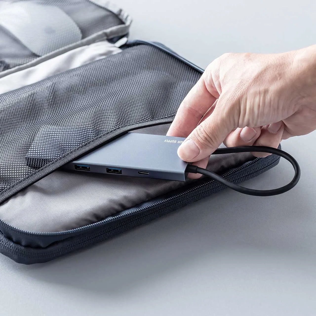手が複数のポートを持つグレーのUSBハブを、メッシュ素材の黒いガジェットポーチに収納している様子