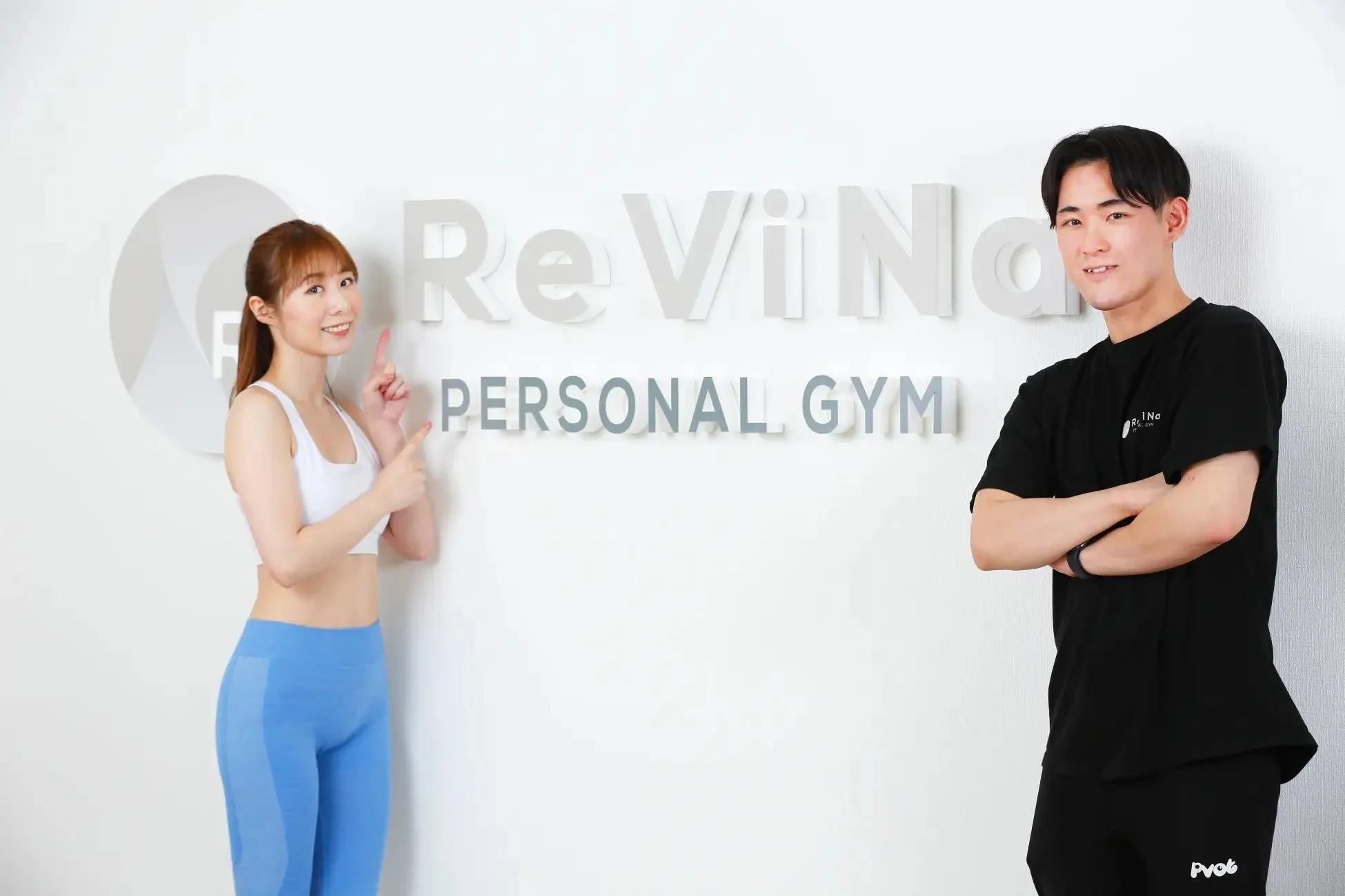 ReViNa PERSONAL GYMのロゴを背景に笑顔で立つトレーナーと女性