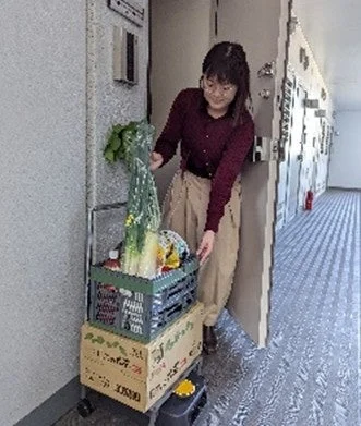 アパートの廊下で、女性がカートに積まれた新鮮な野菜や箱を運んでいる