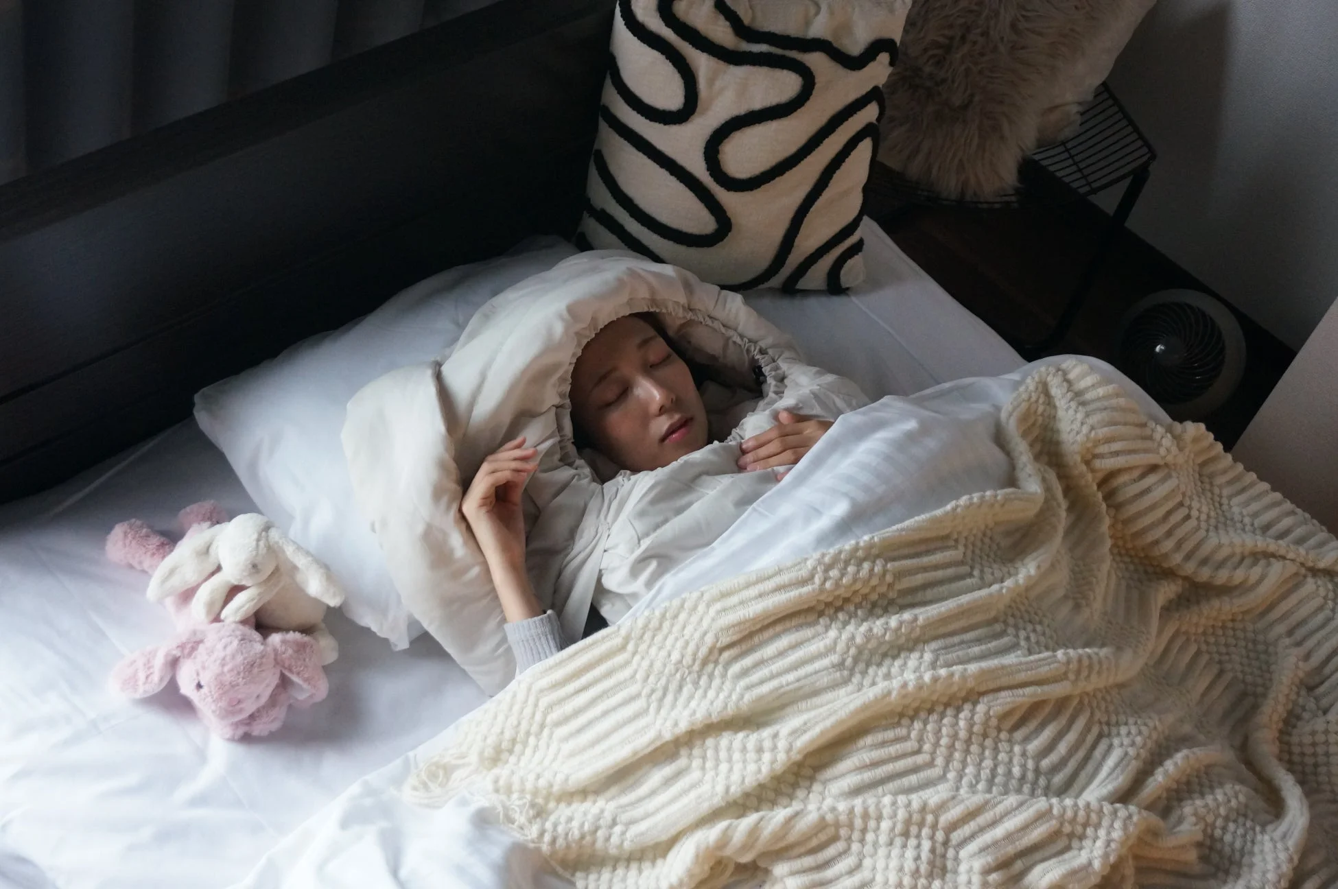白い布団と編み込みのブランケットに包まれてベッドで眠る女性の姿です。枕元には可愛らしい2匹のうさぎのぬいぐるみが寄り添っており、穏やかな休息の時間が流れている様子が伺えます。