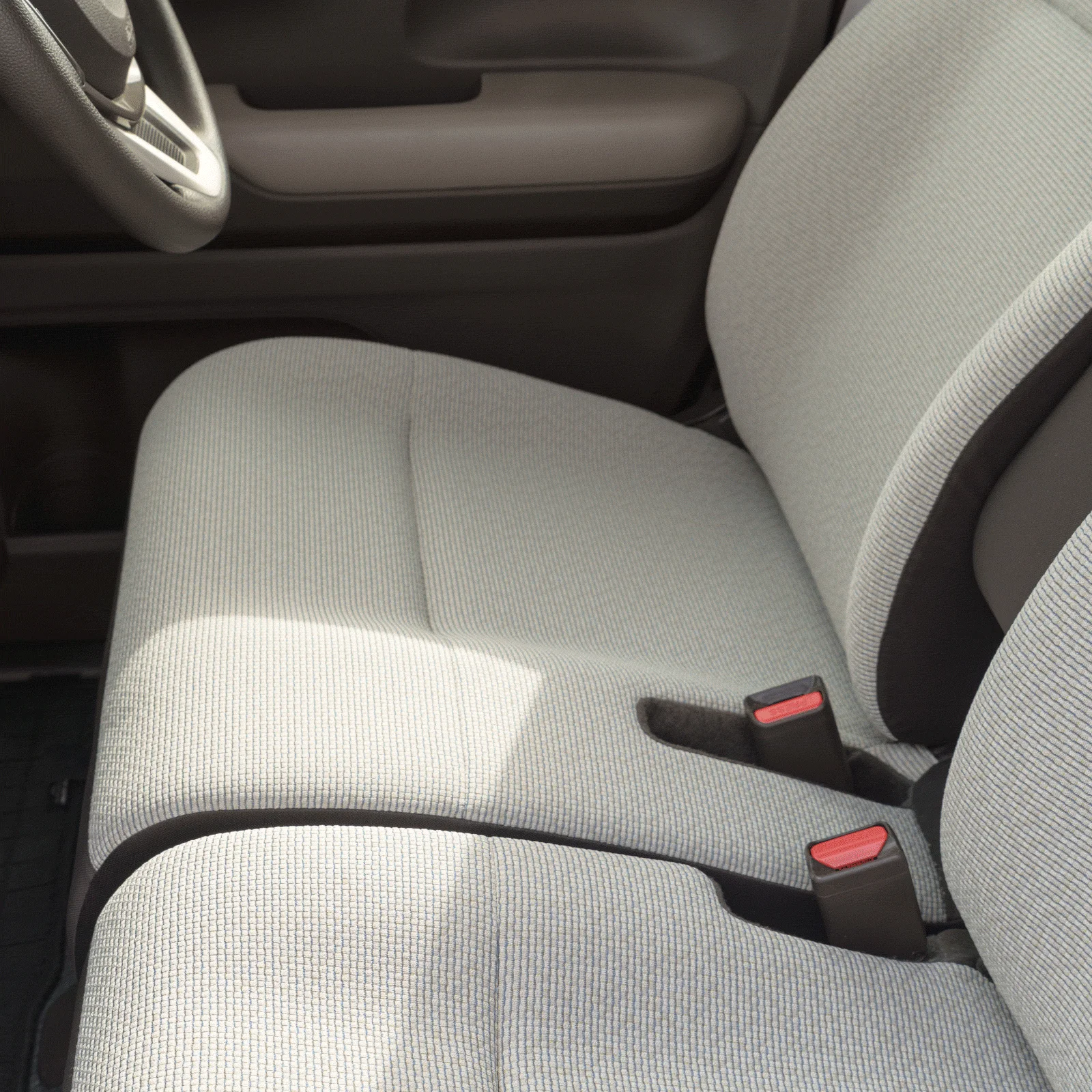 自動車の運転席または助手席の内装を写した画像です。明るいグレーのファブリックシートが特徴で、2つのシートベルトバックルが見えます。ステアリングホイールの一部やドアパネルも写り込んでおり、清潔感のある車内空間が表現されています。