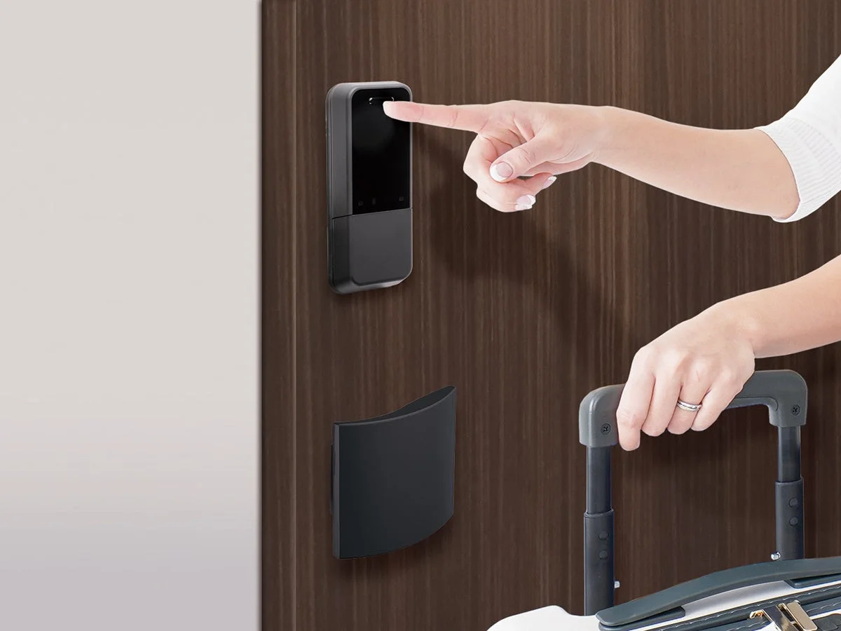 木目のドアに取り付けられたスマートロックに指をかざし、同時にスーツケースのハンドルを握っている人物が写っています