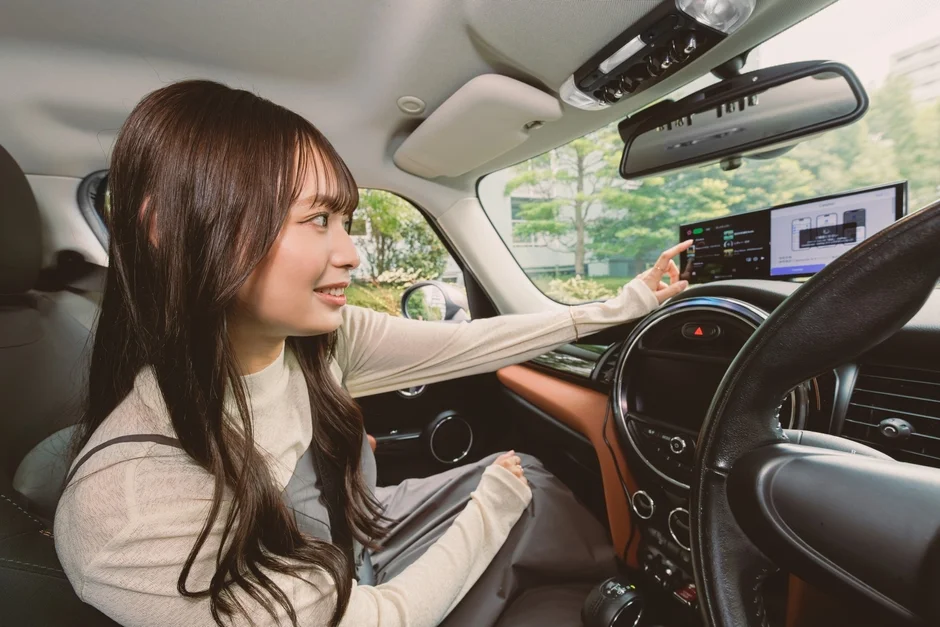 若い女性が車の運転席に座り、中央の大型ディスプレイを操作している様子です。音楽アプリのような画面が表示されており、テクノロジーを活用した現代のカーライフを想起させます。
