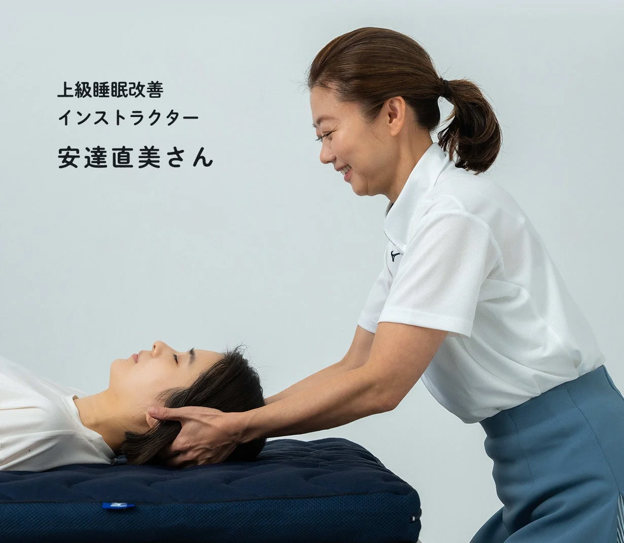 上級睡眠改善インストラクターの安達直美さんが、仰向けに寝ている人の頭部を優しく支え、施術を行っている様子です。快眠をサポートする場面と推測されます。