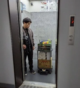 エレベーター内で、男性が業務用スナックや日用品を積んだカートを運んでいる