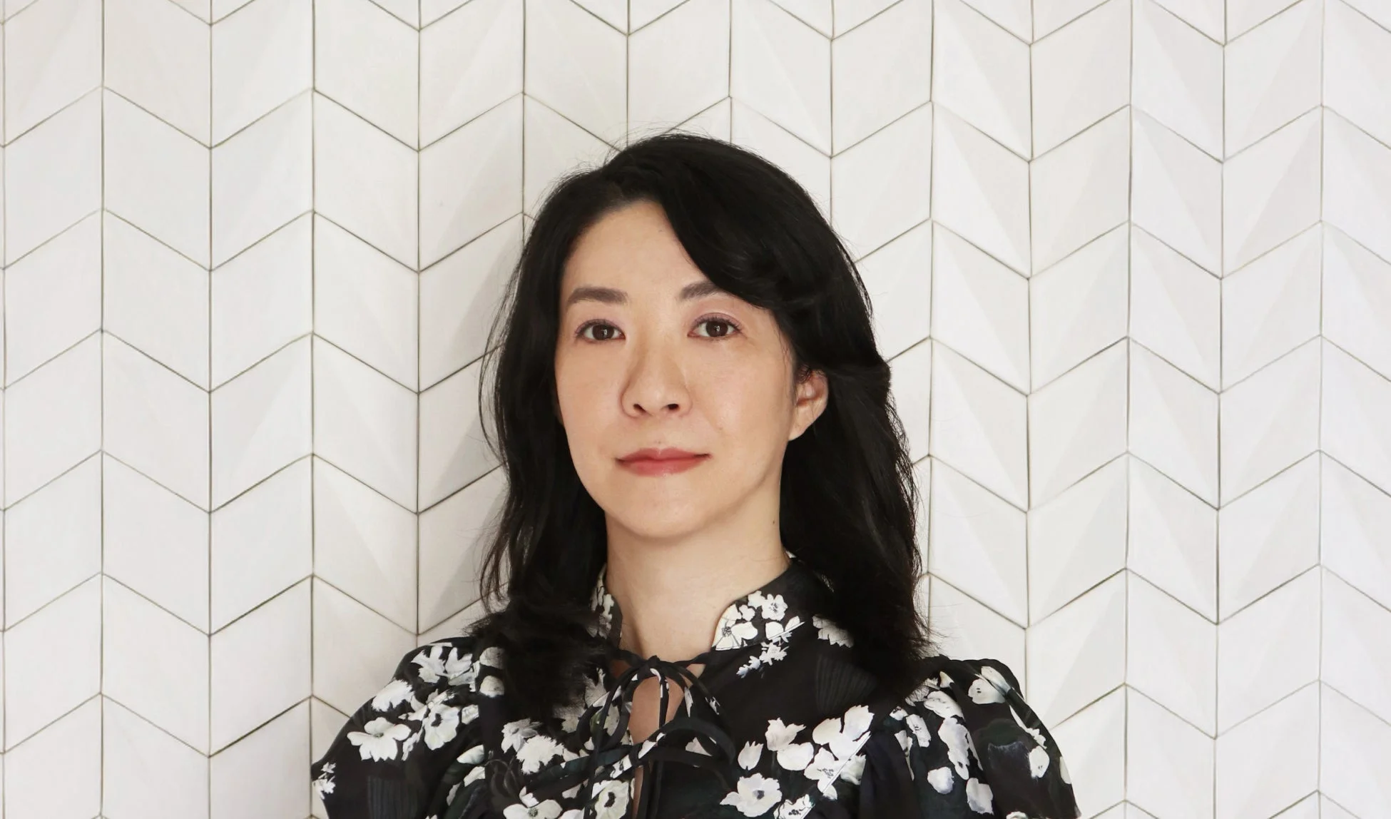 白い幾何学模様の壁を背景に、黒い花柄のトップスを着たアジア人女性のポートレート