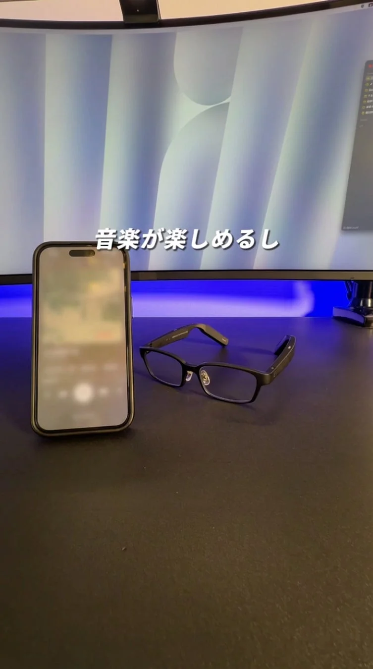 デスク上に置かれたiPhoneとスマートグラスを写した画像です。背景のPCモニターには「音楽が楽しめるし」というテキストが表示されており、スマートグラスで音楽を楽しむ様子を表現しています。