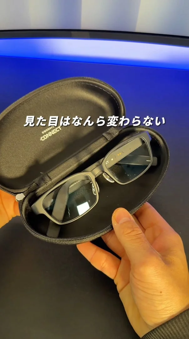 手に持たれた黒いケースに入ったスマートグラスが写っています。画像には「見た目はなんら変わらない」というテキストが表示されており、通常のメガネと変わらないデザインであることを強調しています。