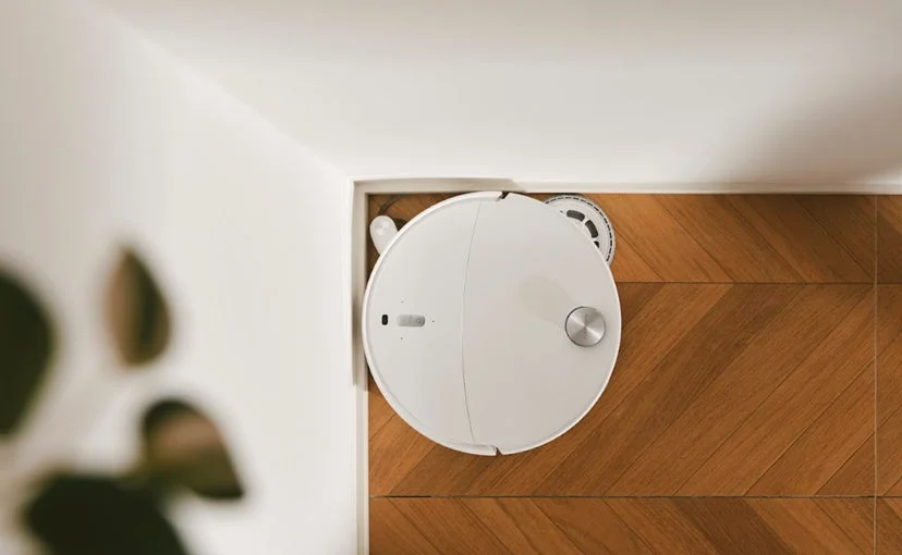 白いロボット掃除機が木目のフローリングの部屋の隅に置かれている様子を上から捉えた画像です。