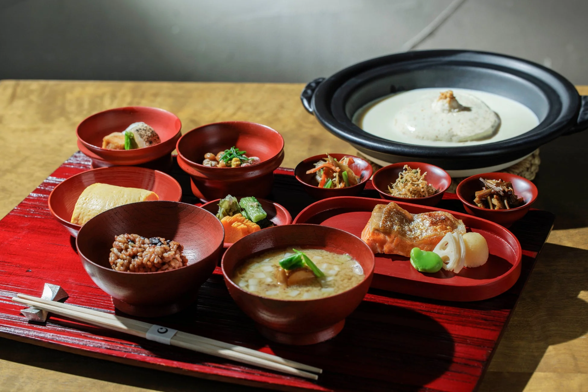 赤いお膳に美しく盛り付けられた豪華な和食の定食です。焼き魚、味噌汁、ご飯、玉子焼き、様々な小鉢に加え、中央には土鍋に入ったクリーミーな料理が並び、旅館や料亭で提供されるような、彩り豊かでバランスの取れた食事が食欲をそそります。
