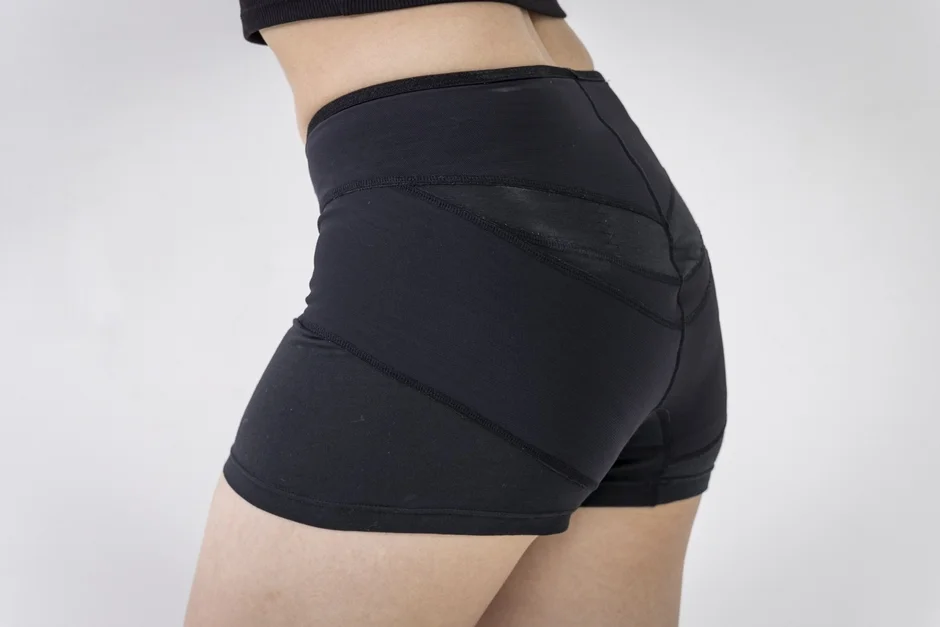 白い背景の前で、黒いスポーツショートパンツを着用した女性の下半身が写っています。ショートパンツは体にフィットし、デザイン性のある縫い目と部分的に異なる質感の素材が見られます。