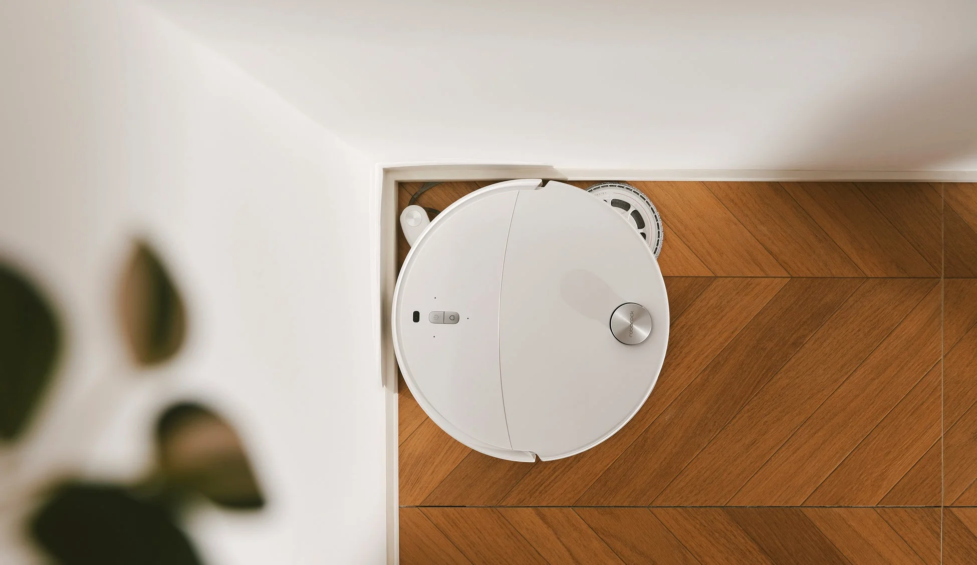 白い丸型のロボット掃除機が、ヘリンボーン柄の木製フローリングの上を掃除している様子を上から捉えた画像です。壁際に設置されており、スマートな室内風景を表現しています。